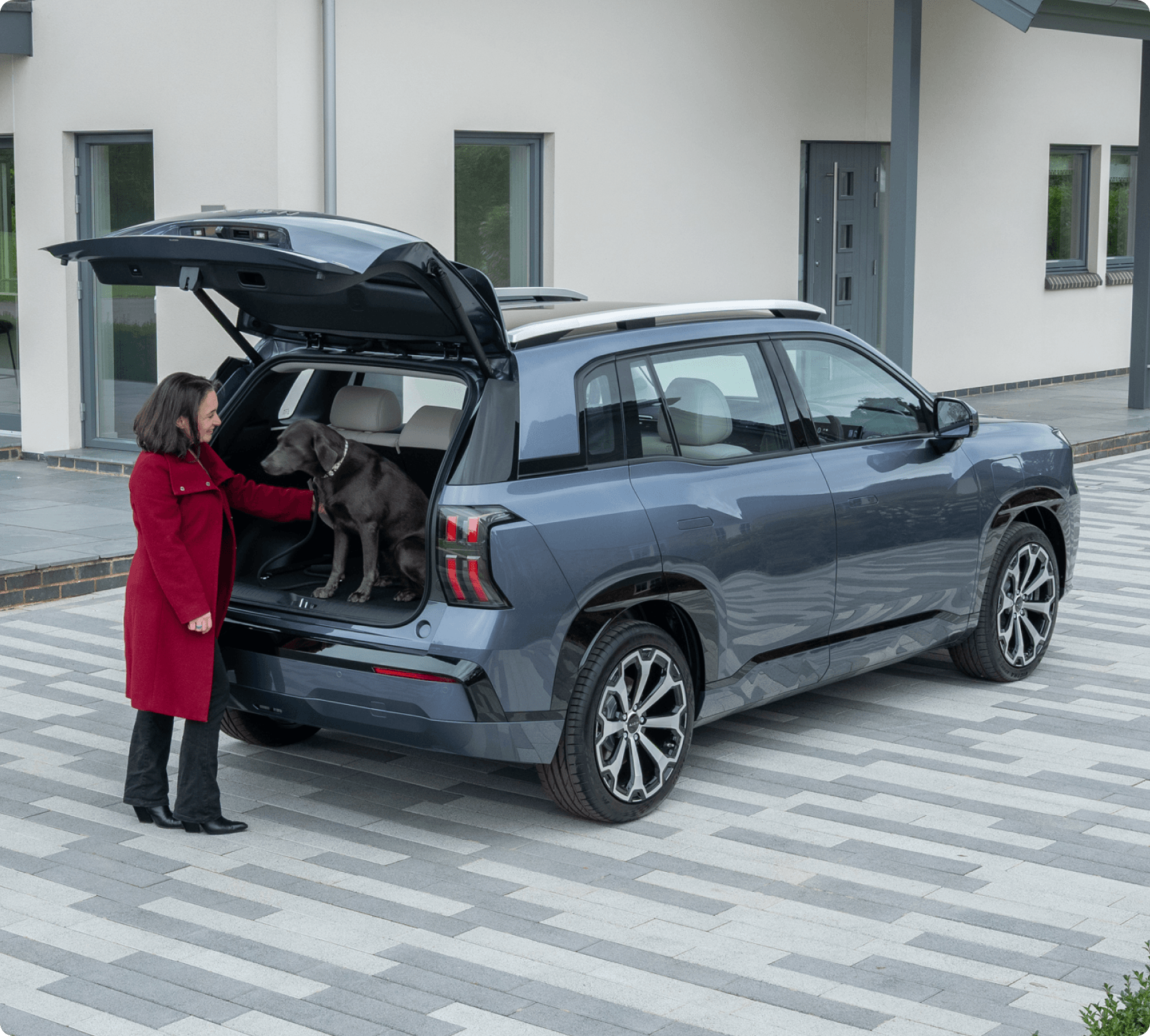Woman in a red coat helping a dog out of the boot of a AION V parked on a driveway outside a modern home