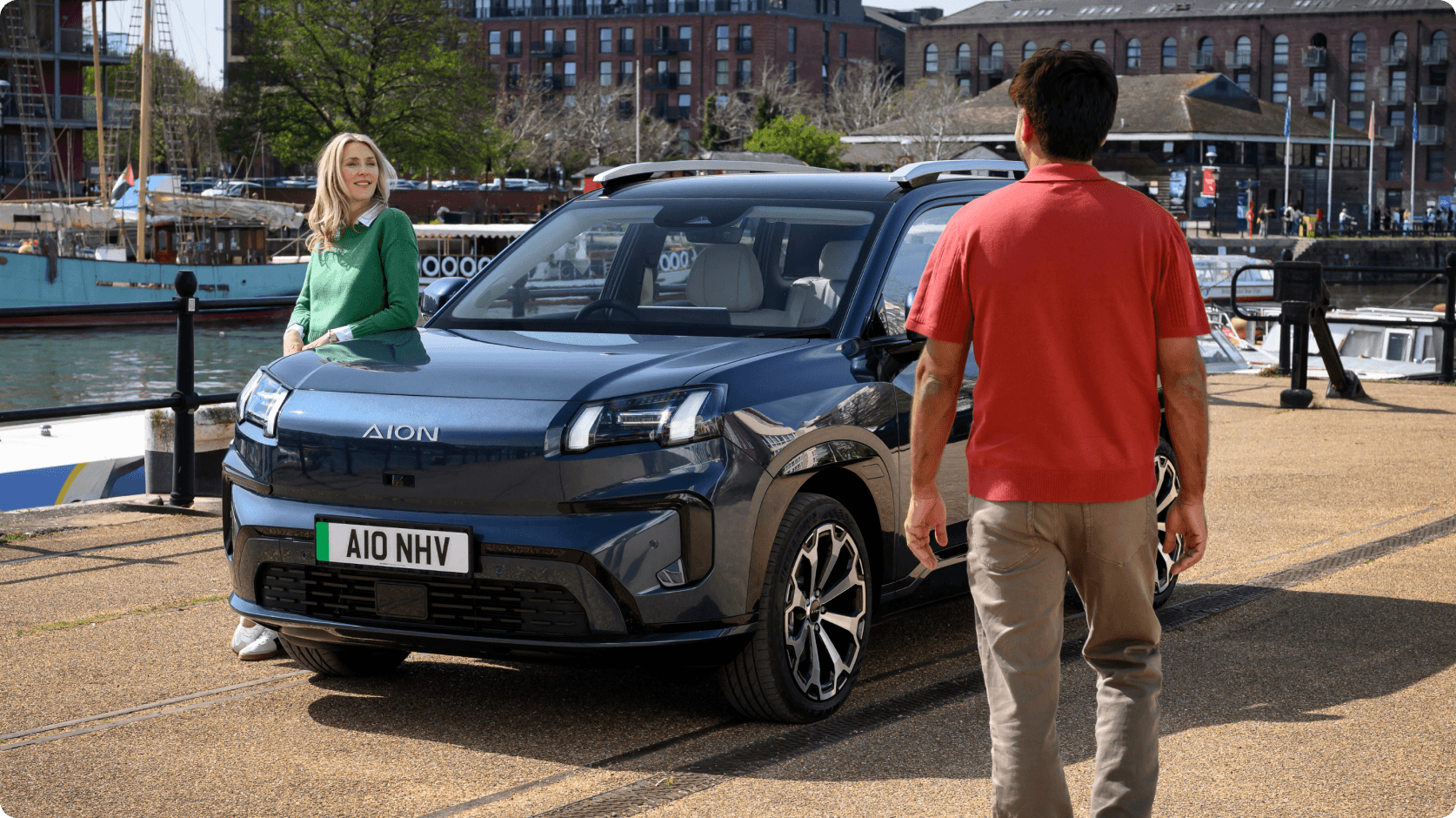 AION V parked by a marina as two people stand beside the vehicle on a waterfront promenade